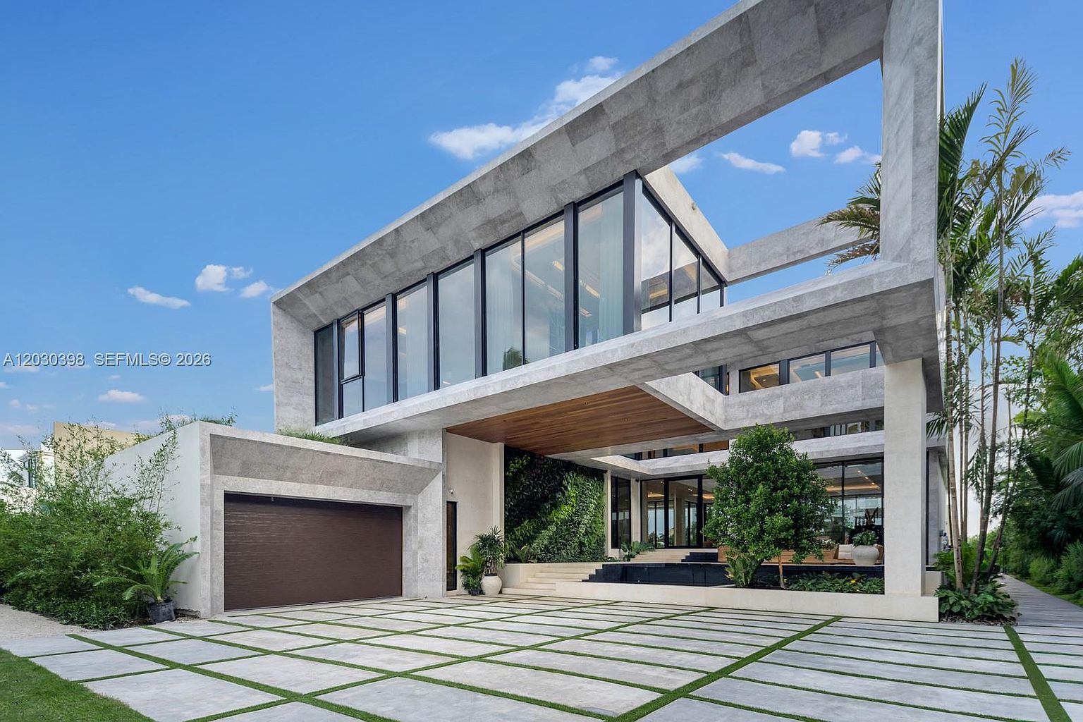 This striking modern residence features a bold, geometric architectural design with extensive use of concrete and large glass panels. The front facade includes a spacious driveway with integrated grass pavers, a prominent garage, and a covered entryway accented by a warm wood-paneled ceiling. A lush vertical garden wall adds a vibrant organic element to the sleek, contemporary structure.