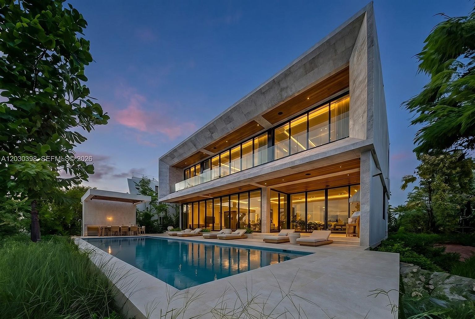 This striking modern residence features a minimalist architectural design with clean lines and a prominent concrete frame. The home showcases extensive floor-to-ceiling glass walls that open onto a spacious patio area, which includes a rectangular swimming pool and several lounge chairs. The scene is captured at dusk, with warm interior lighting creating a welcoming contrast against the cool blue tones of the twilight sky.