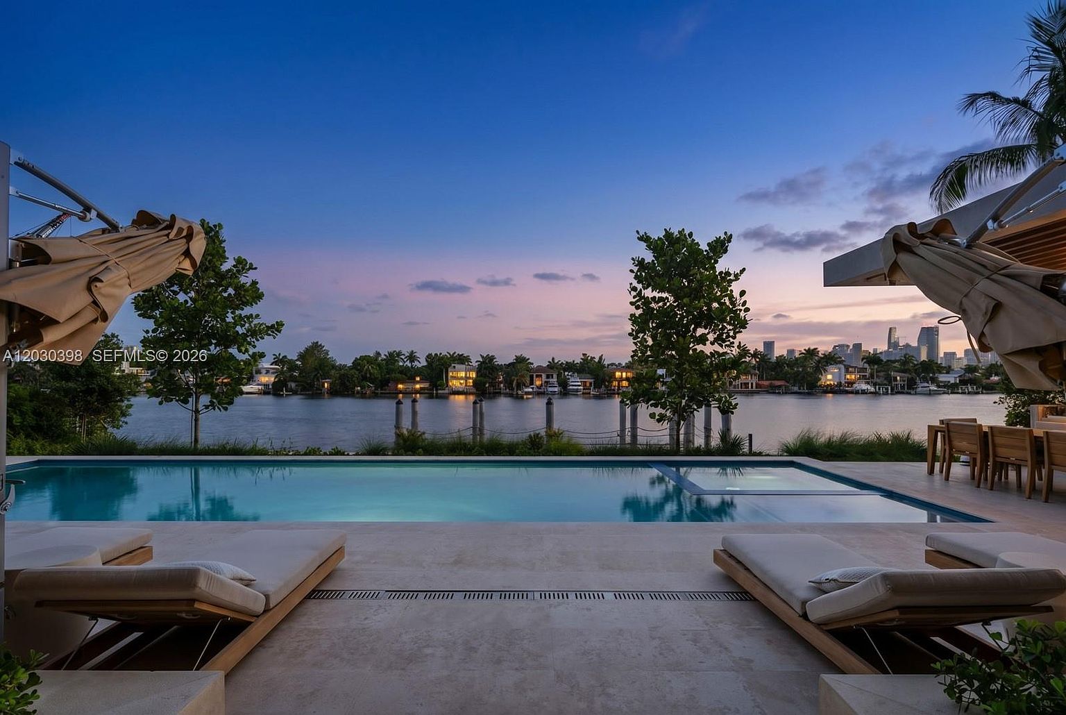 This luxurious waterfront pool deck offers a serene, cinematic perspective looking out over a calm canal toward a distant city skyline at dusk. The foreground features two sleek lounge chairs positioned on a light-toned stone patio, leading directly to a rectangular infinity-edge pool and integrated spa. The scene is framed by two large umbrellas and lush greenery, creating an inviting and sophisticated outdoor living space.