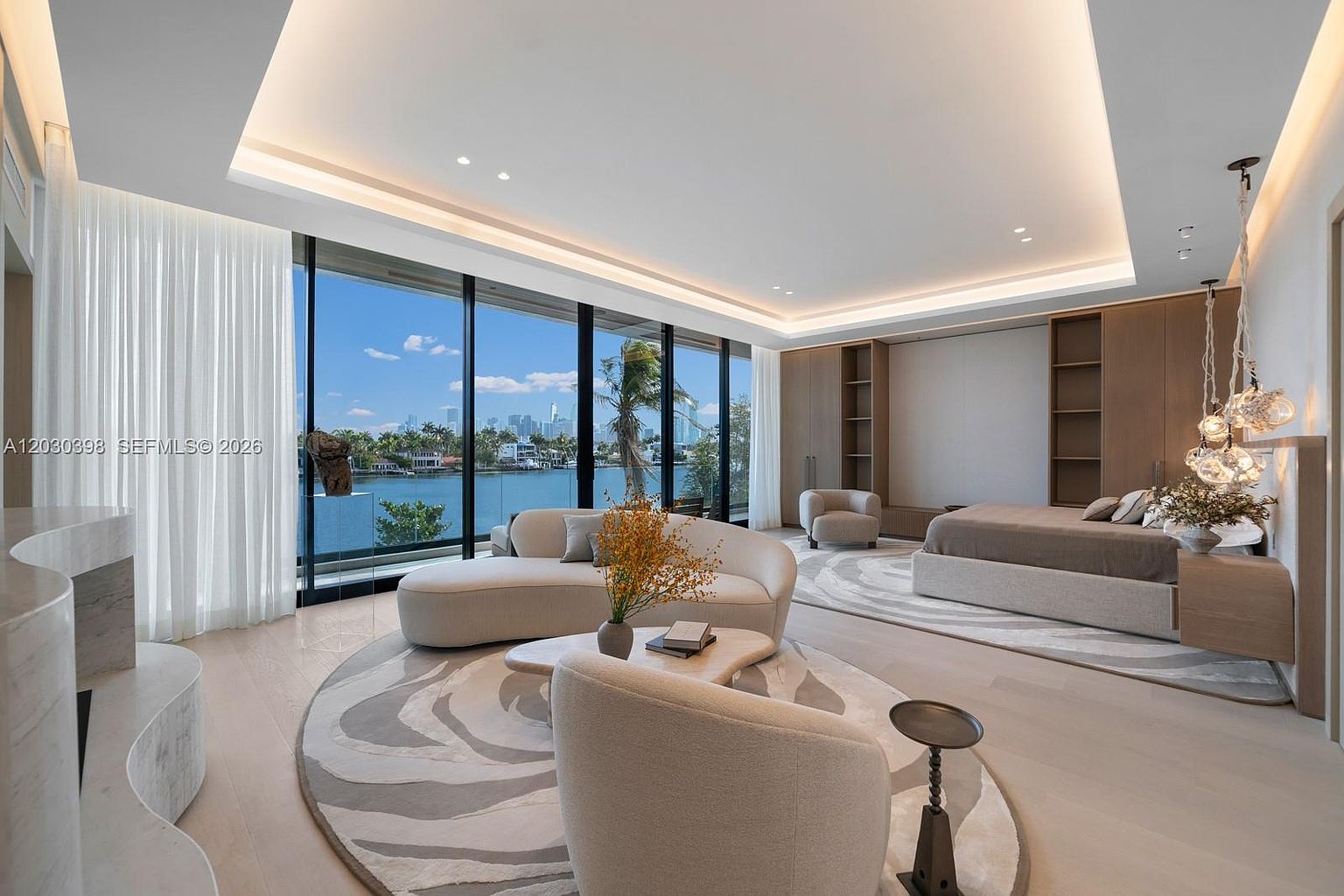 This expansive primary bedroom suite features a seamless blend of sleeping and lounge areas, anchored by a large, neutral-toned bed and a curved sofa. The room is defined by floor-to-ceiling windows that provide an unobstructed, panoramic view of the waterfront and city skyline. A sophisticated, layered lighting design, including recessed ceiling lights and decorative glass pendants, enhances the serene and luxurious atmosphere of the space.