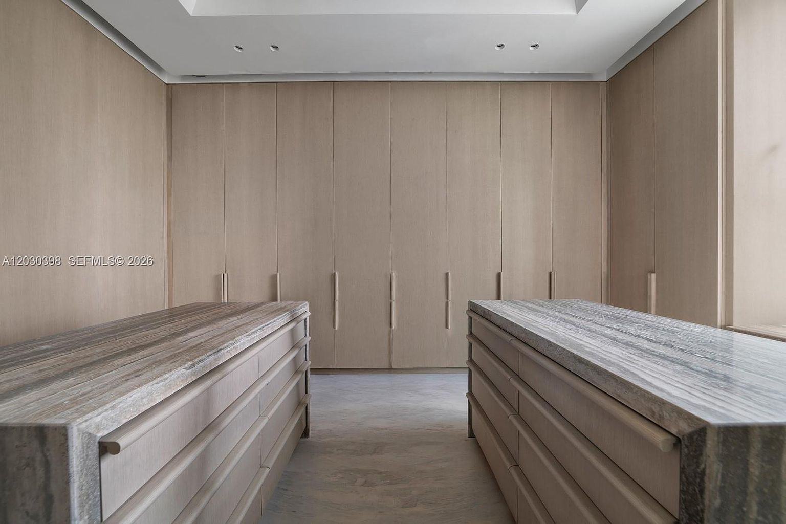 This sophisticated walk-in closet features a symmetrical layout with two large central islands topped with patterned stone surfaces. The walls are lined with extensive light wood cabinetry, providing ample storage behind clean, handle-less doors. The space is illuminated by recessed ceiling lights, creating a bright, minimalist, and highly organized atmosphere.
