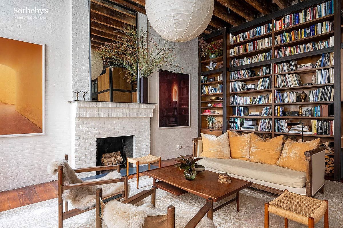 This sophisticated living room features a striking white-painted brick fireplace as a focal point, complemented by a large, floor-to-ceiling mirror and a massive paper lantern pendant light. A wall-to-wall wooden bookshelf filled with books adds warmth and character, while the seating area is anchored by a plush sofa with ochre pillows and mid-century modern armchairs draped in sheepskin. The room exudes a cozy, intellectual, and curated atmosphere, blending industrial elements like exposed ceiling beams with comfortable, textured furnishings.