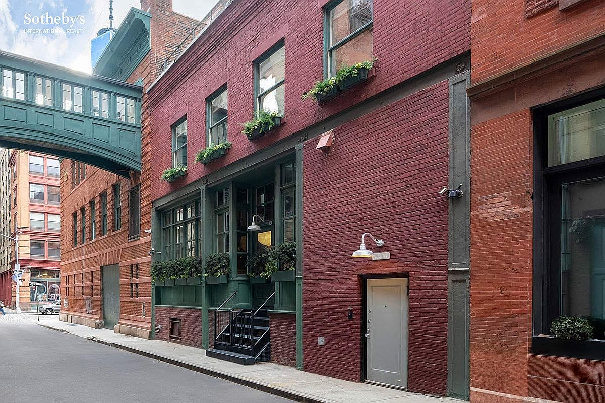 This street-level view captures the charming, industrial-chic facade of a red brick building in an urban setting. The structure features dark green window frames, matching trim, and a distinctive elevated walkway connecting to an adjacent building, all accented by lush window boxes. The perspective is a low-angle shot from the street, emphasizing the building's historic character and unique architectural details.