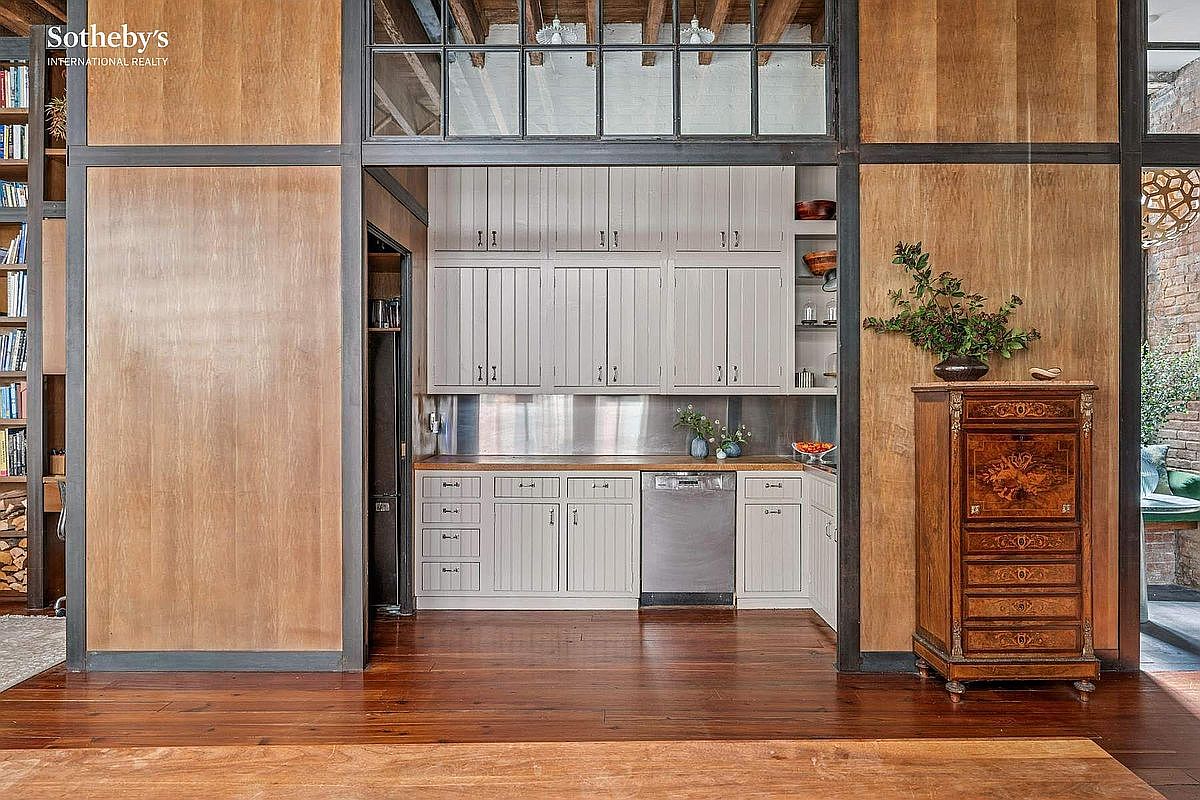 This kitchen features a rustic-industrial aesthetic, characterized by warm wood-paneled walls and a dark metal frame surrounding the cooking area. The space includes light grey cabinetry, a stainless steel dishwasher, and a wooden countertop, creating a cozy yet functional culinary nook. The perspective is a straight-on, eye-level shot that highlights the contrast between the modern appliances and the vintage-inspired wooden furniture nearby.