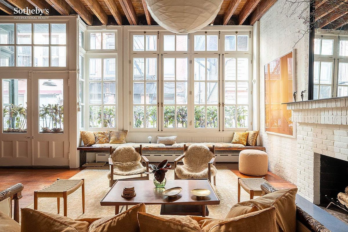 This bright and airy living room features a large, multi-pane window wall that floods the space with natural light, highlighting the warm wood flooring and exposed ceiling beams. The room is furnished with cozy, textured seating, including two sheepskin-covered chairs and a built-in window bench, centered around a low-profile wooden coffee table. A white-painted brick fireplace adds a touch of rustic charm, creating a sophisticated yet inviting atmosphere perfect for relaxation.