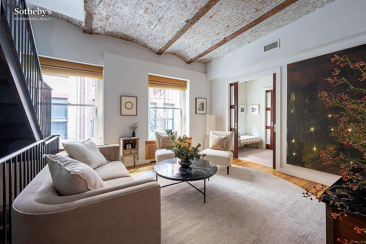 This elegant living room features a unique vaulted brick ceiling and warm hardwood floors, creating a sophisticated and inviting atmosphere. The space is furnished with a plush sofa, two armchairs, and a central marble coffee table, all arranged on a soft area rug. A large, dark abstract painting serves as a dramatic focal point, while a doorway leads into an adjacent room, enhancing the open and airy feel of the home.