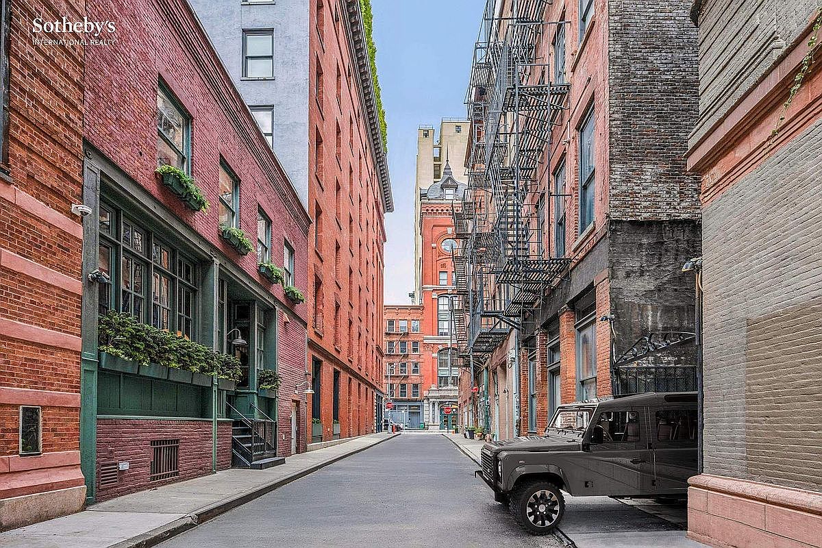 This perspective captures a charming, narrow cobblestone-style street in a historic urban neighborhood, framed by classic red brick buildings. A rugged, dark-colored SUV is parked on the right, while the left side features a building with elegant green-trimmed windows and lush window boxes. The scene evokes a sophisticated, cinematic atmosphere typical of high-end city living.