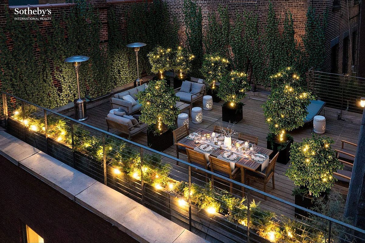 This high-angle, cinematic shot captures a sophisticated rooftop terrace at dusk, illuminated by warm string lights and glowing potted trees. The space features a well-appointed dining area and a comfortable lounge section, all set against a backdrop of lush greenery and brick walls. The elegant lighting and thoughtful layout create an inviting, luxurious atmosphere perfect for evening entertaining.