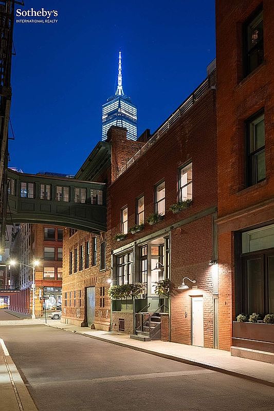 This striking exterior view captures a historic brick building at twilight, featuring a unique elevated skybridge connecting to an adjacent structure. The scene is anchored by the iconic, illuminated spire of the One World Trade Center rising in the background, creating a dramatic contrast between classic architecture and modern city landmarks. The warm glow of exterior wall sconces highlights the textured brick facade and the inviting entryway, evoking a sophisticated urban atmosphere.