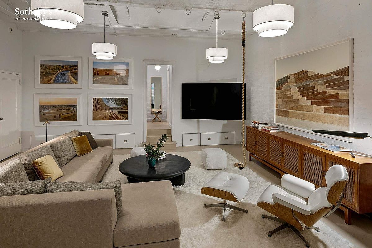 This sophisticated living room features a bright, airy aesthetic with high ceilings, white brick walls, and a plush, light-colored area rug. The space is furnished with a large, comfortable sectional sofa, a classic Eames lounge chair, and a mid-century modern wooden credenza, all centered around a sleek black coffee table and a wall-mounted television. A unique rope feature hangs from the ceiling, adding a playful, artistic touch to the clean, contemporary design.