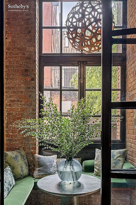 This cozy, light-filled nook features exposed brick walls and a large, multi-paned window that frames an urban view. A circular stone table holds a large glass vase with lush greenery, centered between built-in green cushioned seating adorned with patterned throw pillows. A striking, geometric pendant light hangs above, adding a modern architectural touch to the rustic, intimate space.