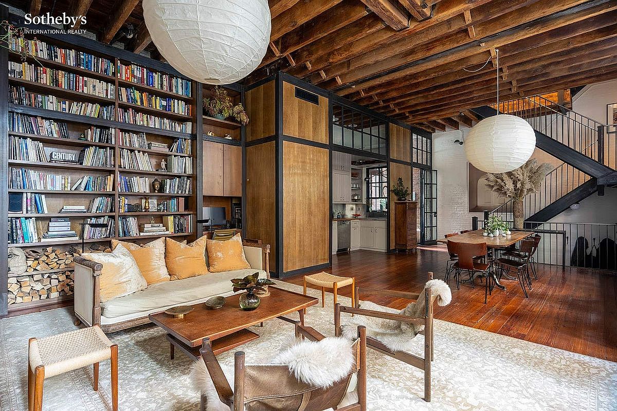 This expansive, open-concept living area features a striking blend of industrial and rustic design, highlighted by exposed wooden ceiling beams and a massive floor-to-ceiling library wall. The space is anchored by a cozy seating area with a plush sofa and fur-accented chairs, while a dining area and staircase are visible in the background. Large paper globe pendant lights add a soft, modern touch to the warm, wood-toned interior, creating an inviting and sophisticated atmosphere.