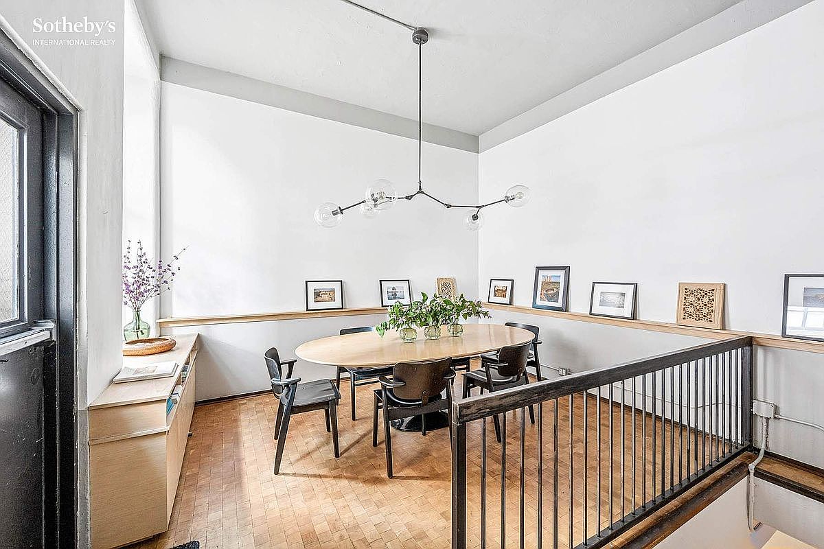 This bright and airy dining area features a modern oval wooden table surrounded by sleek black chairs, positioned beneath a contemporary branching chandelier. The space is defined by light-toned wood flooring, a minimalist sideboard, and a gallery wall of framed art, all overlooking a staircase with a black metal railing. The room offers a sophisticated, open-concept feel with clean lines and a neutral color palette.