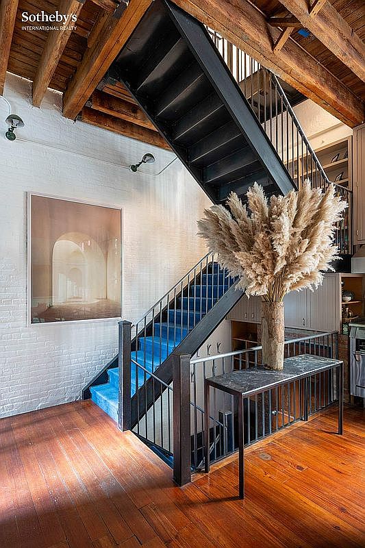 This striking interior space features a dramatic industrial-style staircase with black metal railings and vibrant blue carpeted steps, set against a textured white brick wall. A large, minimalist framed print hangs on the wall, while a tall vase filled with pampas grass sits atop a sleek, dark marble-topped console table in the foreground. The warm wooden flooring and exposed ceiling beams create a sophisticated, loft-like atmosphere that blends rustic charm with modern design.
