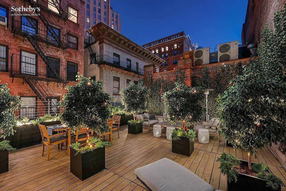 This sophisticated urban rooftop terrace features warm wooden decking, lush potted trees, and comfortable outdoor seating arrangements, creating an inviting oasis amidst the city skyline. The space is framed by classic brick architecture and fire escapes, offering a blend of industrial charm and refined relaxation. The perspective is a wide-angle shot that captures the entire terrace, emphasizing its potential as a private, high-end entertainment area.