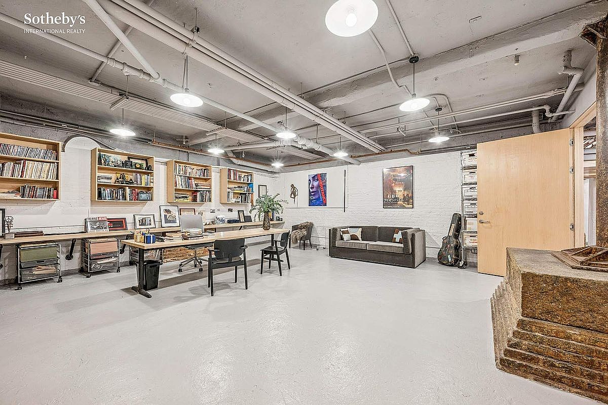 This spacious, industrial-style home office features exposed ceiling pipes, white-painted brick walls, and a polished concrete floor. The room is furnished with a long wooden desk, multiple bookshelves, a comfortable sofa, and modern lighting fixtures, creating a creative and functional workspace. The wide-angle perspective highlights the open layout and the blend of raw architectural elements with professional office amenities.