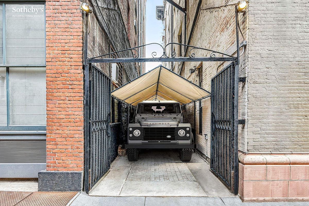 This image captures a unique, private urban parking space nestled between two historic brick buildings. A rugged Land Rover Defender is parked securely under a custom-fitted canvas canopy, framed by ornate black wrought-iron gates that provide a sense of exclusivity and security. The perspective is a straight-on, eye-level shot that emphasizes the narrow, industrial-chic character of this rare city amenity.