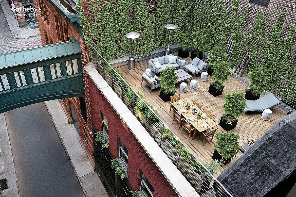 This high-angle aerial perspective showcases a sophisticated urban rooftop terrace featuring a spacious wooden deck, a dining area, and a comfortable lounge section. The space is elegantly framed by lush climbing ivy on the brick walls, potted greenery, and a modern metal railing, creating a private oasis above the street. A unique enclosed pedestrian bridge connects the building to an adjacent structure, adding a distinct architectural character to this luxury outdoor living space.