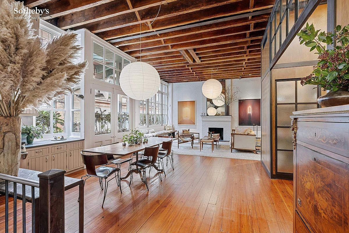 This expansive, open-concept living and dining area features high, exposed wood-beam ceilings and warm hardwood flooring that creates a sophisticated, industrial-chic atmosphere. A long wooden dining table sits prominently in the foreground, while the background reveals a bright, airy living space centered around a white brick fireplace. Large, multi-paned windows flood the room with natural light, highlighting the elegant paper pendant lights and curated art pieces.