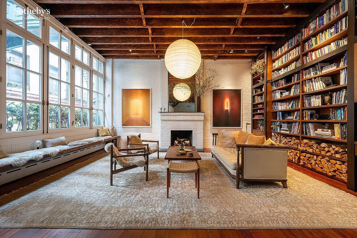 This sophisticated living room features a warm, rustic aesthetic with exposed wooden ceiling beams, a white brick fireplace, and extensive floor-to-ceiling bookshelves filled with volumes. A large, neutral-toned area rug anchors the space, which is furnished with a comfortable sofa, mid-century modern armchairs, and a low-profile coffee table. The room is bathed in natural light from a wall of large, multi-paned windows, creating an inviting and airy atmosphere perfect for relaxation or reading.