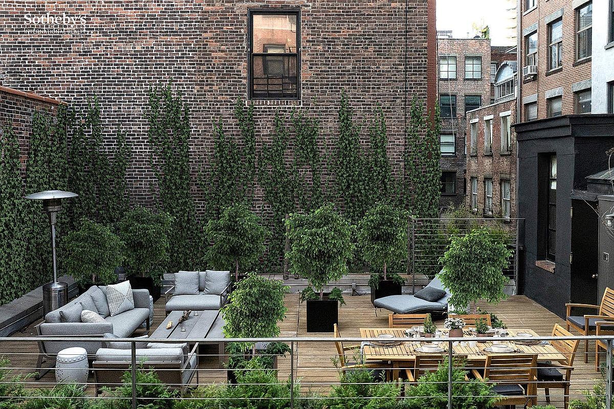 This sophisticated urban rooftop terrace features a spacious wooden deck furnished with a comfortable grey sectional sofa, a dining set, and a lounge chair. The space is framed by a lush living wall of tall, manicured hedges and potted trees, creating a private oasis against a classic brick building backdrop. The elevated perspective captures a serene, high-end outdoor living area perfect for entertaining in a city setting.