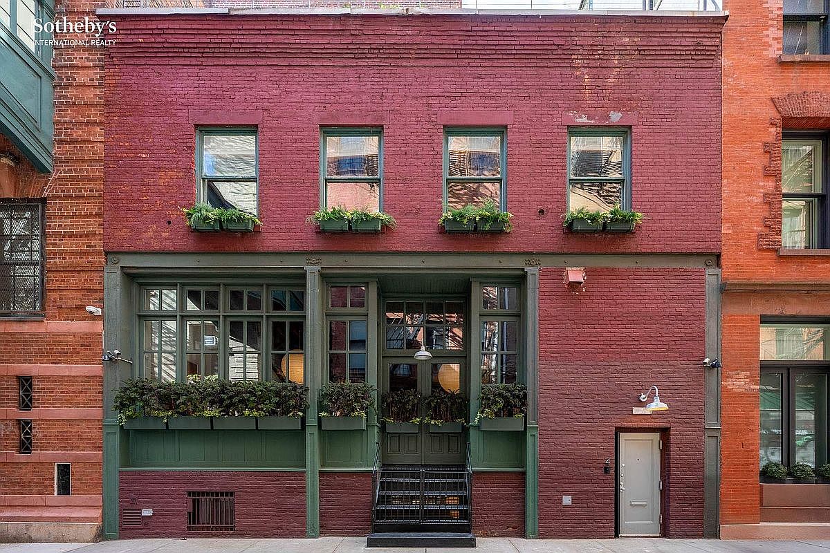 This striking townhouse facade features a deep red brick exterior accented by dark green window frames, doors, and trim. The front entrance is elevated by a short set of stairs, flanked by lush window boxes and large, multi-paned windows that create a sophisticated, urban aesthetic. The composition is captured from a straight-on, eye-level perspective, emphasizing the symmetrical design and the charming, historic character of the building.