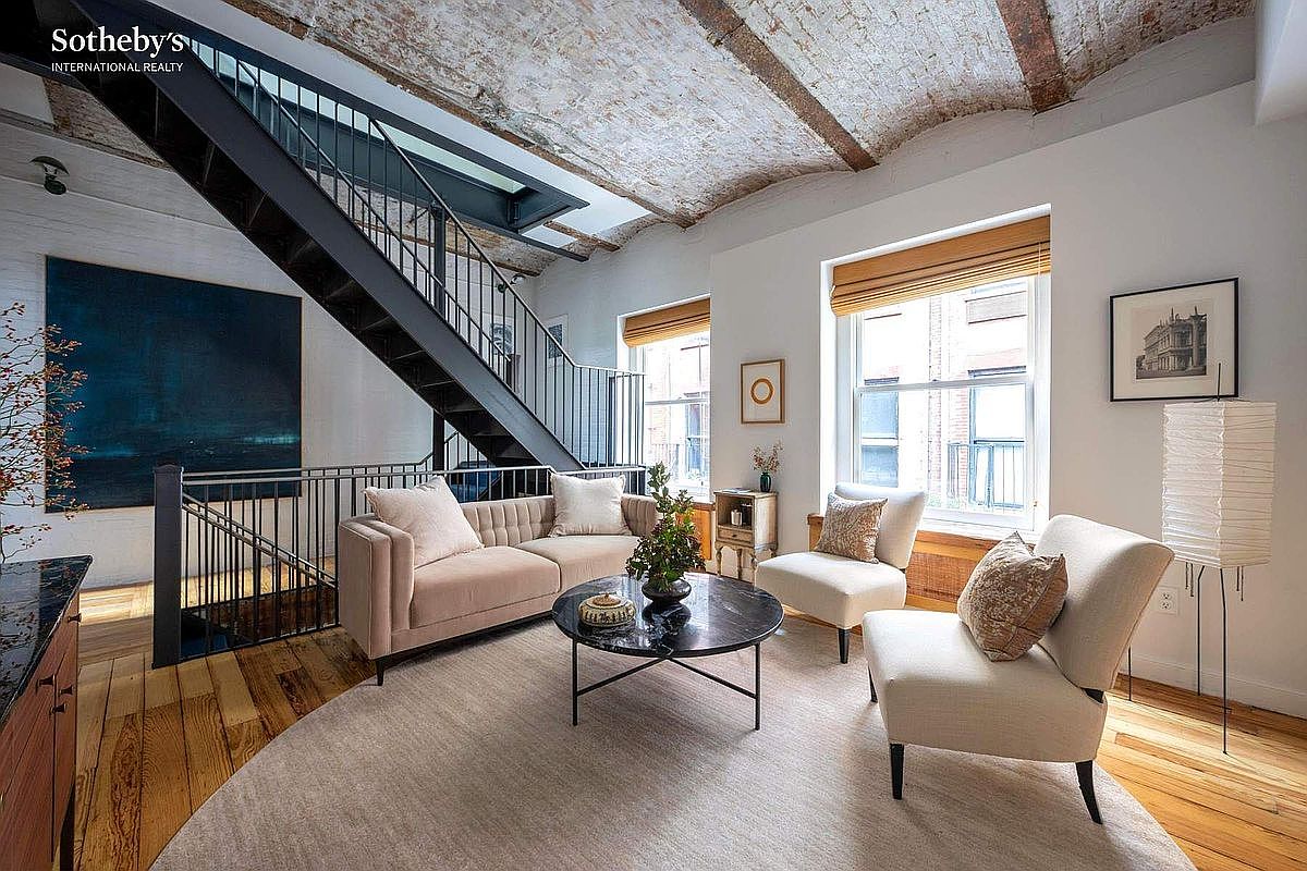 This sophisticated living room features a striking industrial aesthetic, highlighted by a vaulted brick ceiling and a dramatic black metal staircase. The space is furnished with a plush, light-toned sofa and two matching armchairs arranged around a sleek, circular black marble coffee table. Large windows with wooden blinds allow natural light to fill the room, creating an airy and elegant atmosphere that blends historic architectural charm with modern comfort.