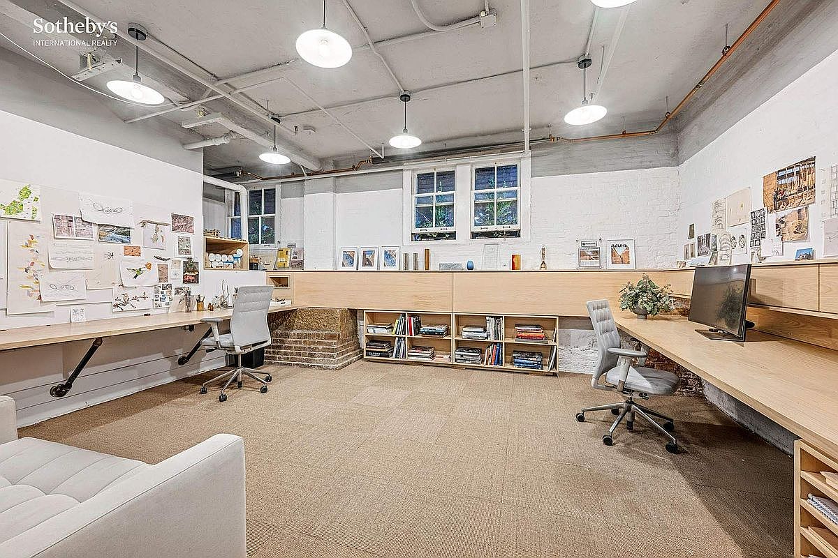 This spacious, industrial-style home office features a long, custom-built wooden desk spanning two walls, providing ample workspace for multiple users. The room is accented by exposed ceiling pipes, a brick foundation element, and large windows that bring in natural light, creating a creative and productive atmosphere. The neutral color palette and organized shelving make it a functional and aesthetically pleasing workspace.