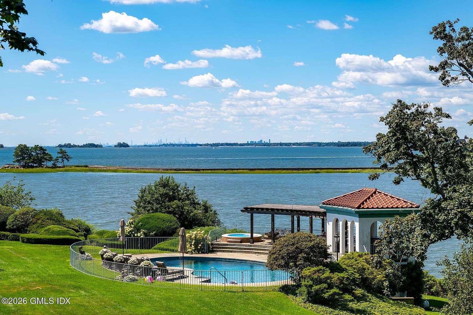 This elevated view captures a serene backyard oasis featuring a curved swimming pool and an adjacent hot tub set against a stunning waterfront backdrop. A classic pergola provides shade near the pool, while a charming gazebo with a terracotta-tiled roof adds a touch of architectural elegance to the landscape. The expansive lawn leads the eye toward the calm bay, with a distant city skyline visible on the horizon under a bright, cloud-dappled sky.
