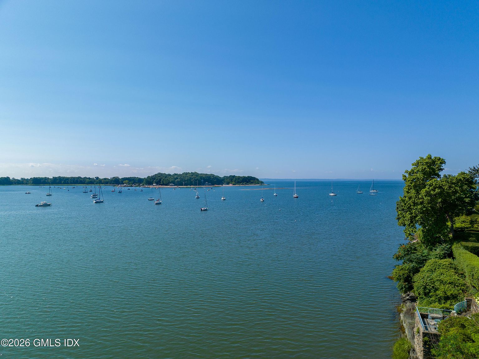 This aerial perspective captures a serene coastal scene featuring a vast, calm body of water dotted with numerous sailboats and small vessels. To the right, a lush, tree-lined shoreline with a small stone-walled terrace provides a grounded foreground element. The clear blue sky and expansive horizon create a tranquil and picturesque atmosphere, highlighting the property's prime waterfront location.