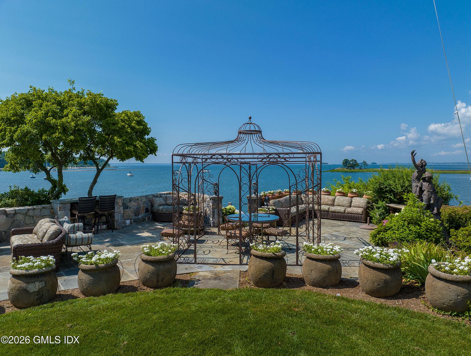This expansive stone patio offers a luxurious outdoor living space overlooking a serene coastal view. The area features a decorative metal gazebo, comfortable wicker-style seating, and numerous stone planters filled with white flowers, all set against a backdrop of the ocean and a clear blue sky. A stone wall borders the patio, and a sculptural statue adds an elegant artistic touch to the landscaped surroundings.