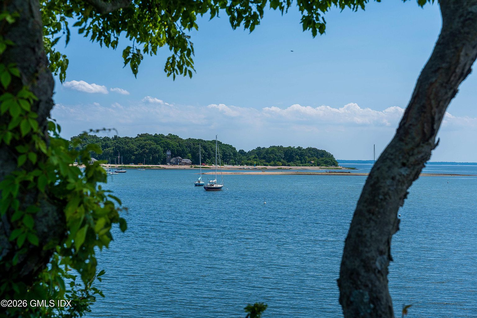 This scenic view captures a tranquil coastal landscape framed by the natural branches of foreground trees. The composition looks out over a calm blue bay toward a distant, tree-lined shoreline featuring a small beach and a prominent building. Several sailboats are anchored in the water, enhancing the serene, nautical atmosphere of this waterfront setting.