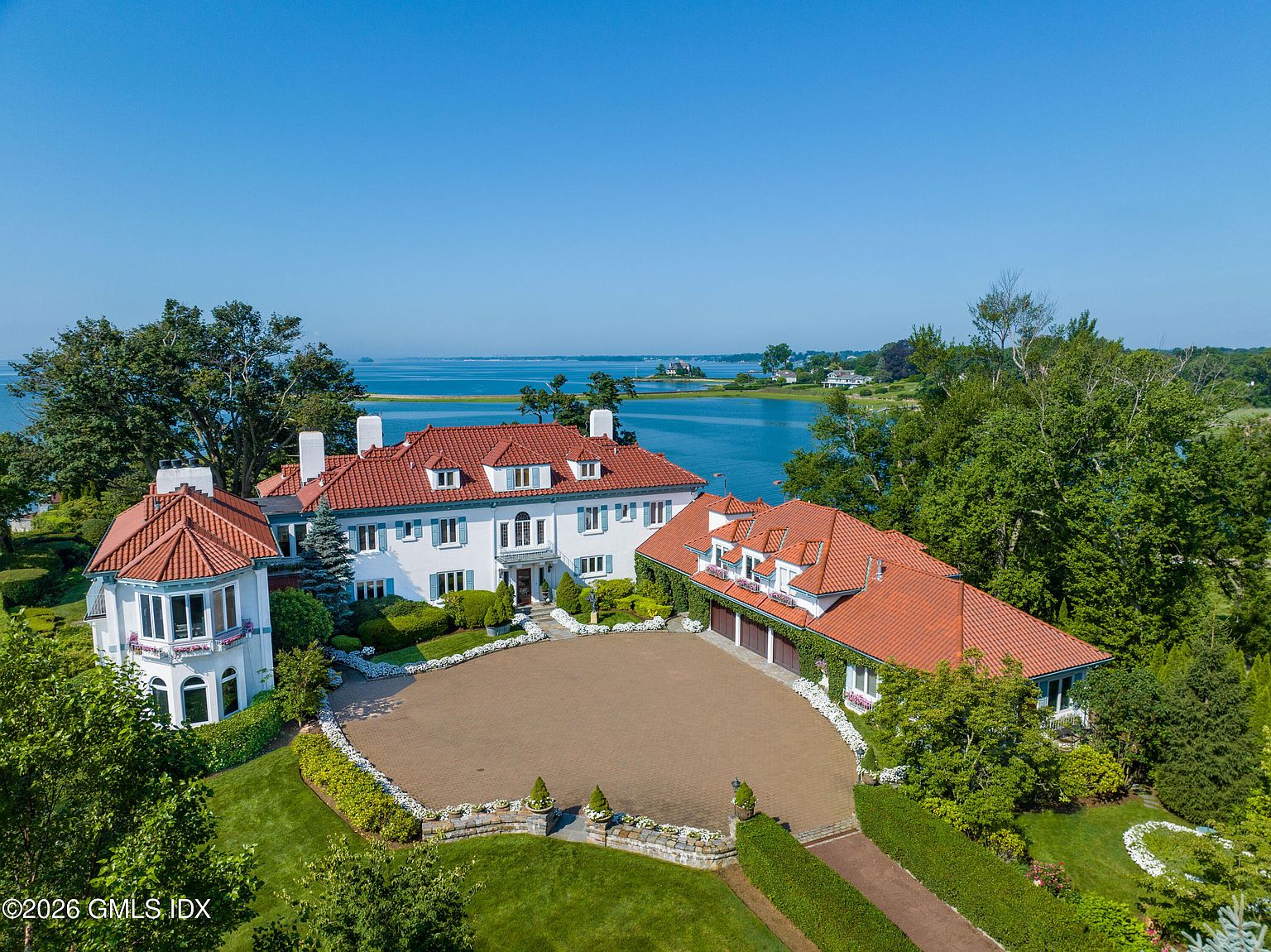 This stunning aerial perspective captures a grand, Mediterranean-inspired estate set against a serene coastal backdrop. The property features a white stucco facade with a distinctive red-tiled roof, a spacious circular paved driveway, and lush, manicured landscaping. The expansive water view and mature trees surrounding the home create a sense of privacy and luxury, highlighting the property's prime waterfront location.