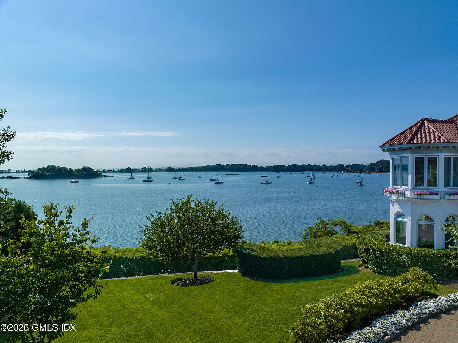 This elevated perspective captures a serene waterfront view from a property, showcasing a lush green lawn, manicured hedges, and a mature tree in the foreground. To the right, a portion of a multi-story home with a red-tiled roof and a balcony with flower boxes is visible. The background features a calm bay filled with numerous sailboats under a clear, expansive blue sky.