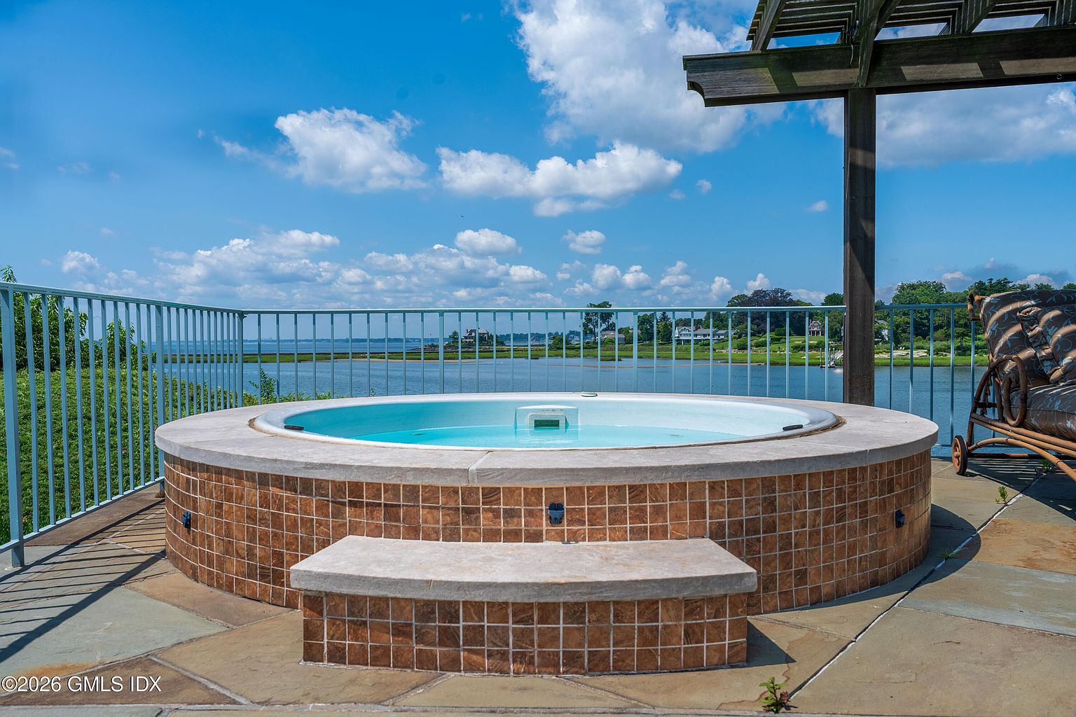 This outdoor space features a circular, tiled spa set into a stone-paved deck, offering a panoramic view of the water and distant shoreline. A metal railing surrounds the perimeter, while a wooden pergola structure provides partial shade to the side. The scene captures a serene, resort-like atmosphere perfect for relaxation.