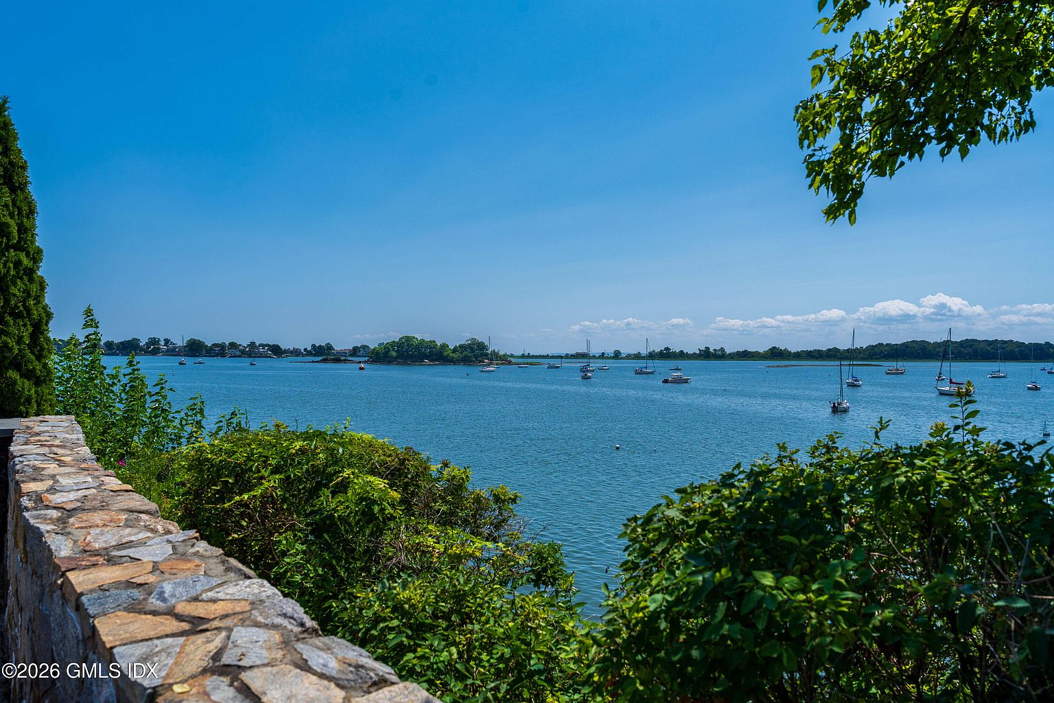 This scenic outdoor view captures a tranquil waterfront setting from a stone-walled vantage point. Lush green foliage and trees frame the foreground, leading the eye toward a calm bay dotted with numerous sailboats under a clear, expansive blue sky. The perspective offers a serene, elevated look at the coastal landscape, highlighting the property's prime location and natural beauty.
