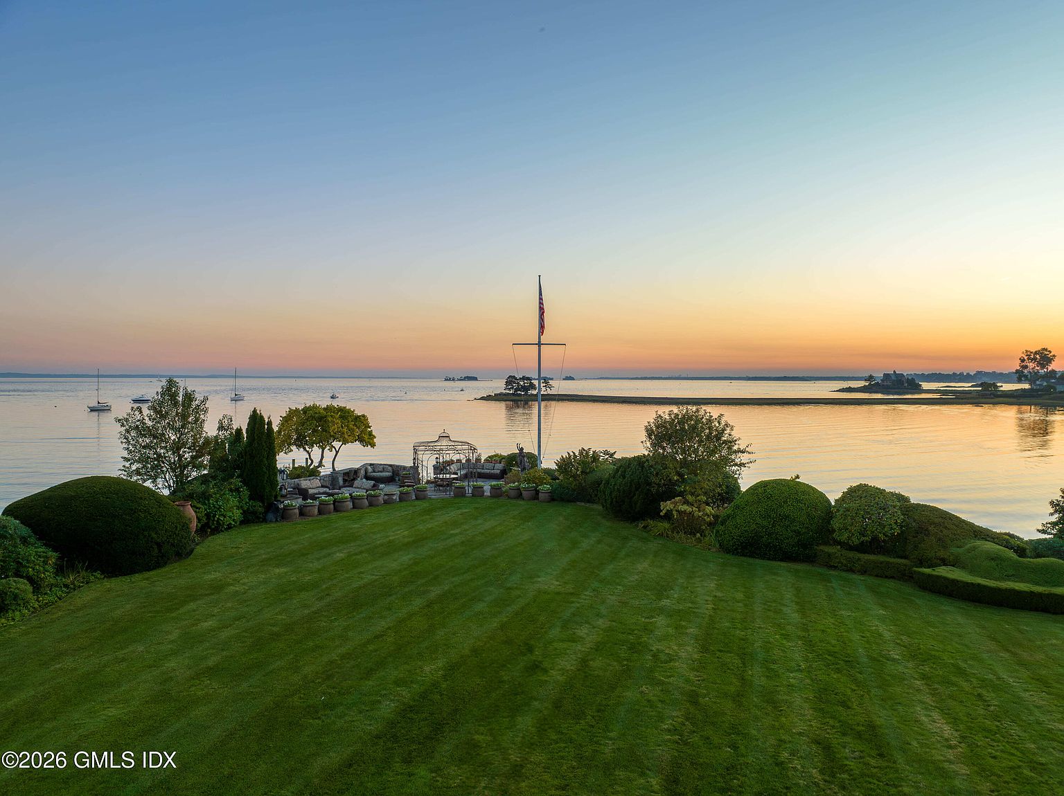 This expansive, manicured lawn overlooks a serene waterfront at sunset, offering a picturesque and tranquil setting. The foreground features lush, sculpted shrubbery and a stone-paved patio area with outdoor seating, while a tall flagpole stands prominently near the water's edge. The horizon is defined by a soft, golden-hued sky reflecting over the calm bay, creating a peaceful and luxurious atmosphere.