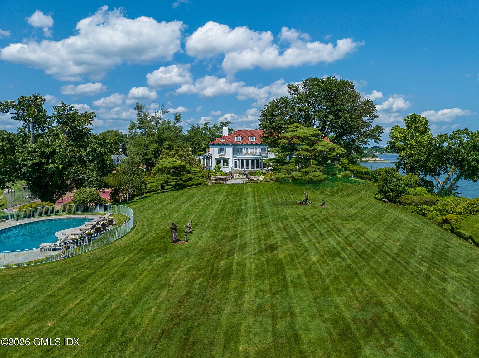 This expansive aerial view captures a grand estate featuring a large, multi-story residence with a distinctive red-tiled roof, set against a backdrop of lush greenery and a serene waterfront. The property boasts a meticulously manicured lawn, a private swimming pool enclosed by a white fence, and several decorative sculptures scattered across the grounds. The perspective provides a cinematic overview of the sprawling landscape, highlighting the seamless integration of luxury architecture with the natural coastal environment.
