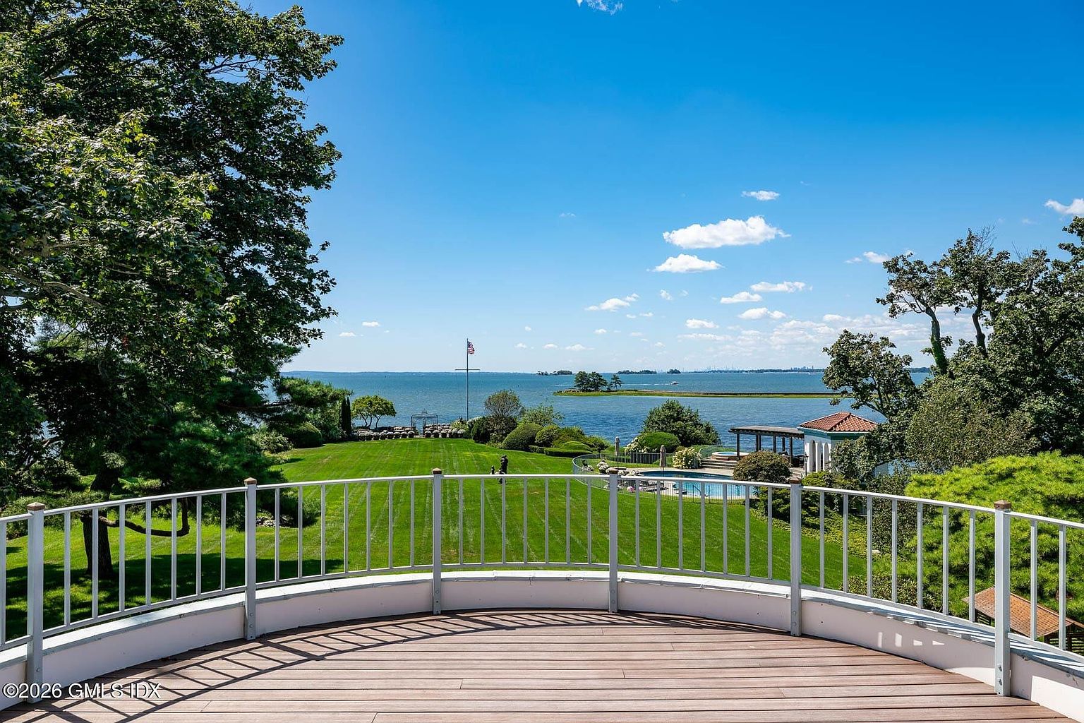 This elevated, curved deck offers a sweeping, panoramic view of a lush green lawn leading down to a serene waterfront. The scene features a clear blue sky, a swimming pool nestled near the water's edge, and mature trees framing the expansive coastal landscape. The perspective is from a high vantage point, emphasizing the property's prime location and tranquil outdoor living space.