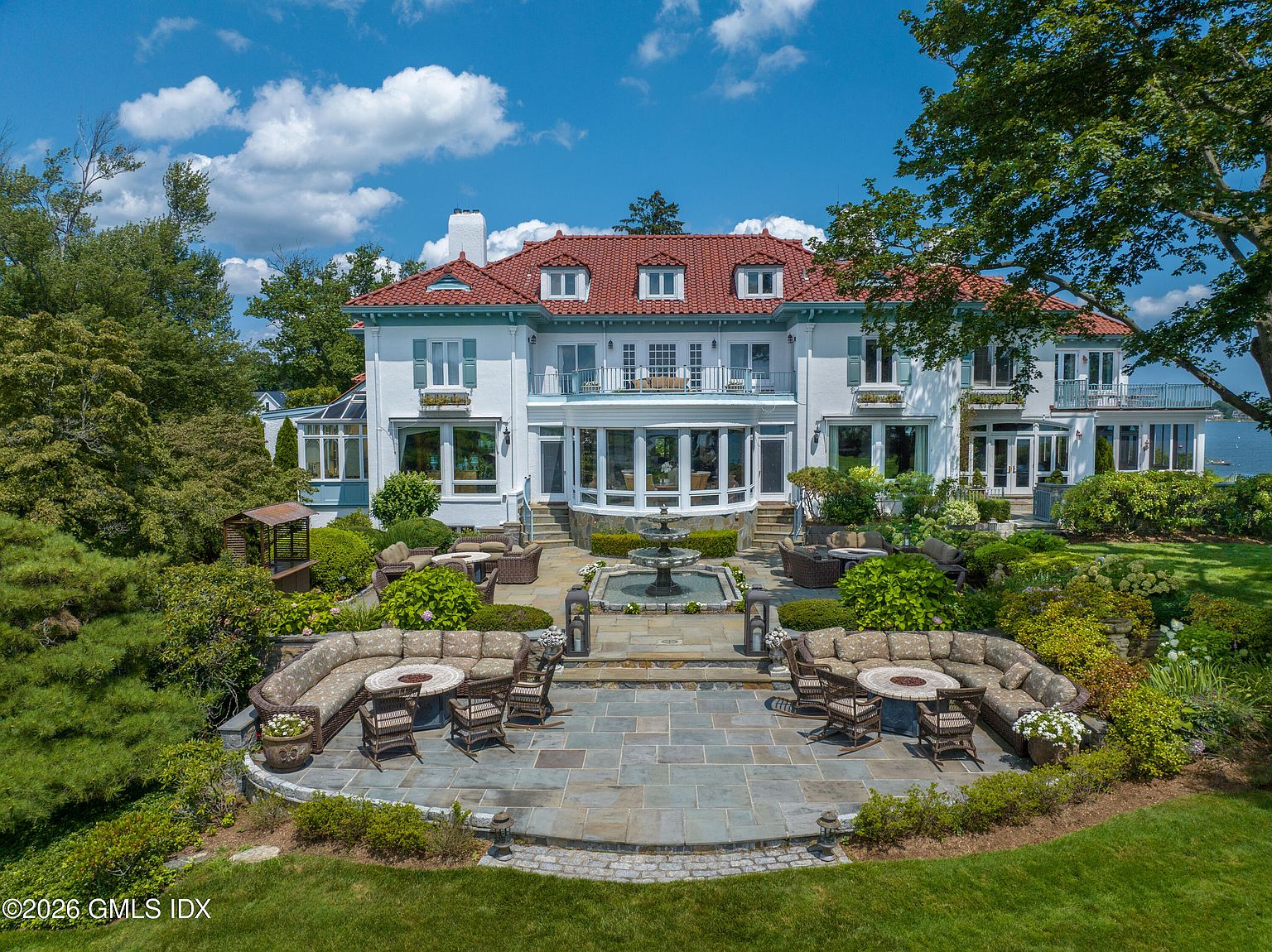 This grand, multi-story estate features a classic Mediterranean-inspired design with a striking red tile roof and crisp white stucco exterior. The rear facade opens onto an expansive stone terrace, complete with a central tiered fountain, multiple curved seating areas with fire pits, and lush, manicured landscaping. Captured from an elevated perspective, the scene highlights the seamless transition between the elegant architecture and the serene, park-like grounds overlooking the water.