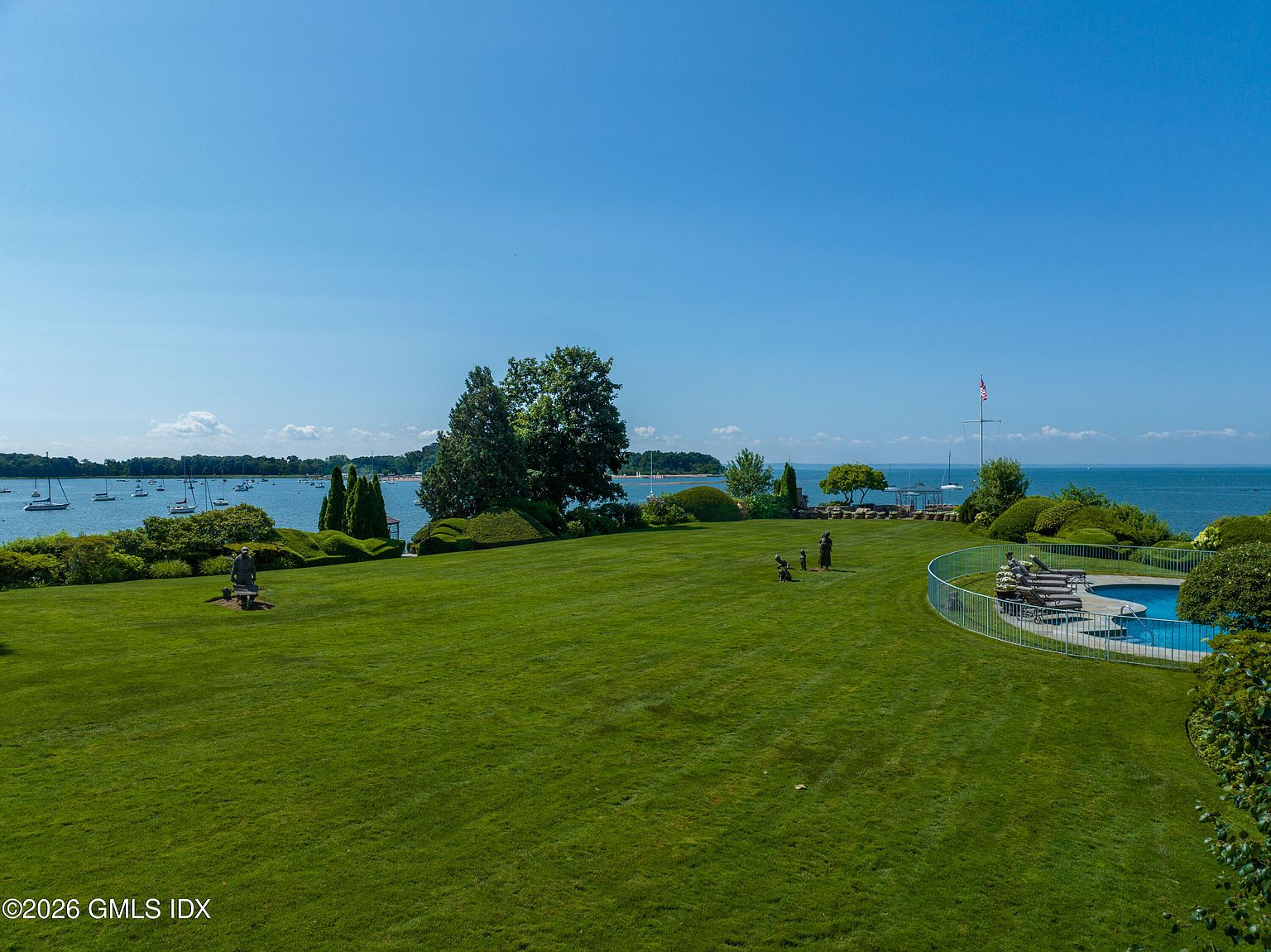 This expansive, manicured lawn overlooks a serene coastal bay filled with sailboats under a clear blue sky. The property features a fenced-in swimming pool area on the right, mature landscaping with sculpted hedges, and decorative statues placed throughout the grounds. The elevated perspective captures the vastness of the waterfront setting, emphasizing the property's prime location and tranquil atmosphere.