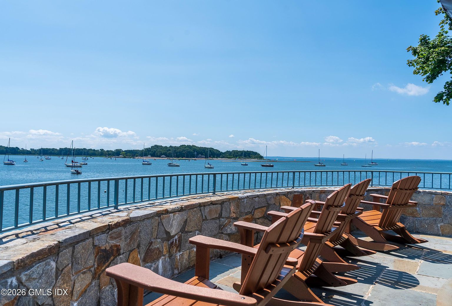 This elevated stone terrace offers a panoramic, cinematic view of a calm bay dotted with numerous sailboats. A row of classic wooden Adirondack chairs is arranged along a sturdy stone wall topped with a metal railing, creating an inviting space for relaxation. The bright, clear sky and expansive water view emphasize the property's prime waterfront location.