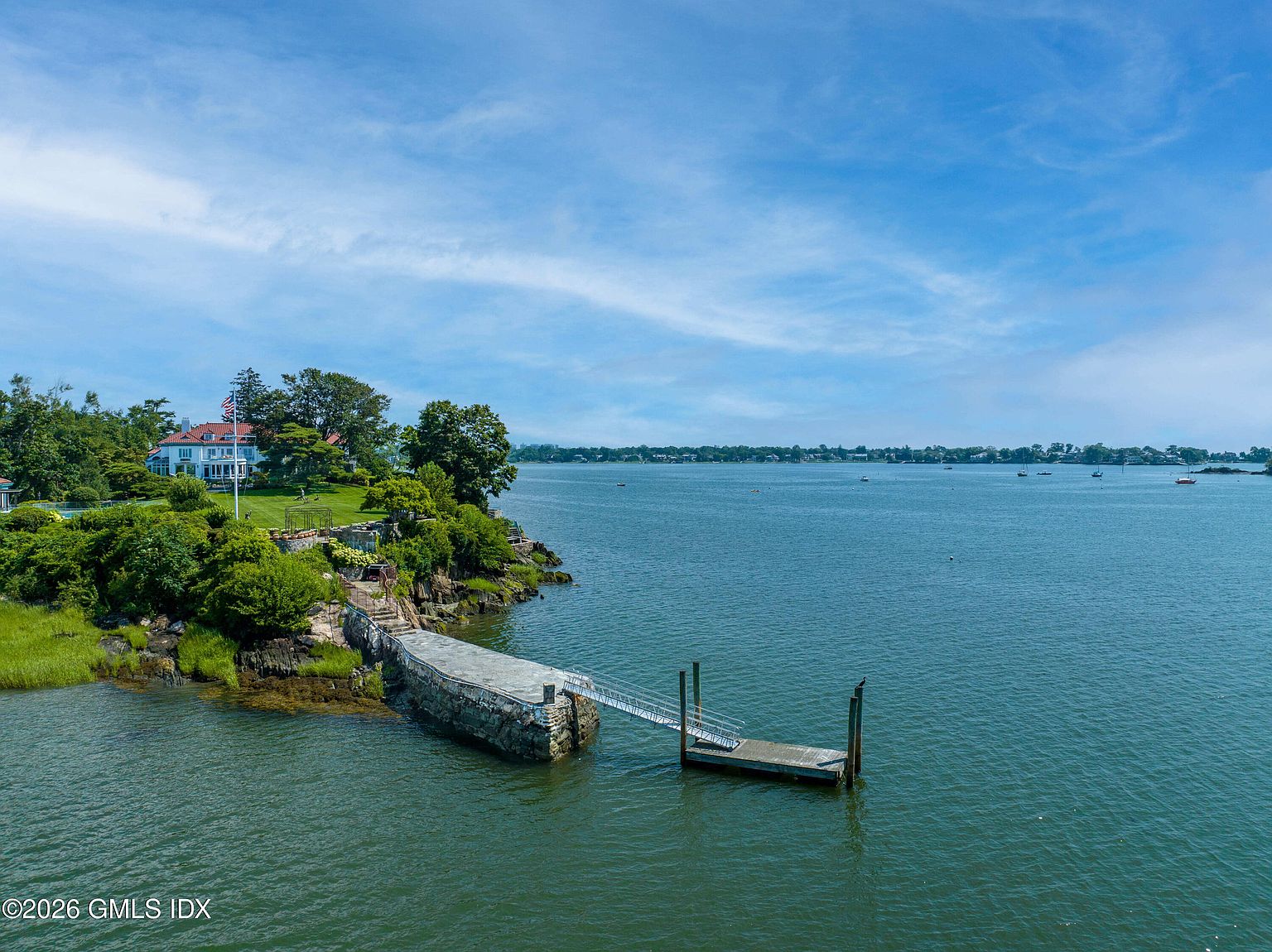 This aerial perspective captures a sprawling waterfront estate situated on a rocky peninsula, showcasing a classic white residence with a red roof nestled among lush greenery. A stone-walled pier extends from the property into the calm, expansive bay, providing private water access. The scene offers a serene and prestigious coastal atmosphere, highlighting the home's prime location and panoramic water views.