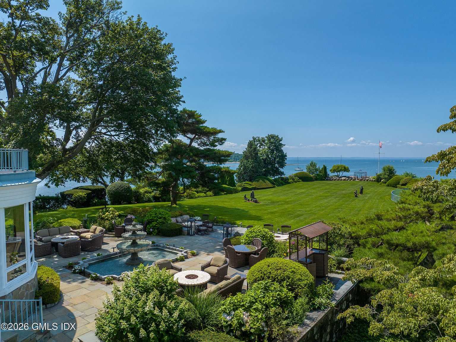 This elevated view captures a sprawling, manicured lawn that extends toward a scenic waterfront horizon. The foreground features a stone-paved patio equipped with multiple seating areas, a tiered water fountain, and fire pits, creating an ideal space for outdoor entertaining. Lush landscaping, mature trees, and a clear blue sky enhance the serene and luxurious atmosphere of this expansive property.