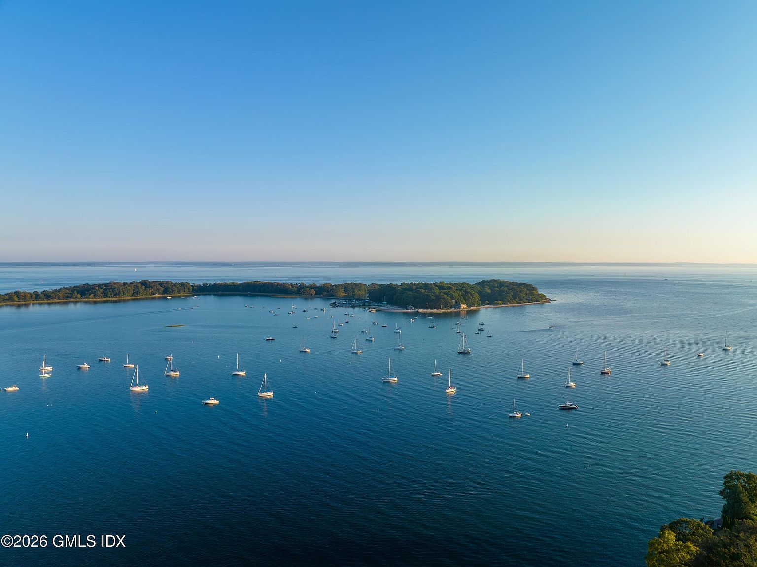 This high-angle aerial view captures a serene coastal landscape featuring a lush, tree-covered island surrounded by calm, deep blue waters. Numerous sailboats are anchored throughout the bay, creating a peaceful maritime atmosphere under a clear, expansive sky. The perspective emphasizes the expansive waterfront setting and the tranquil nature of the location.