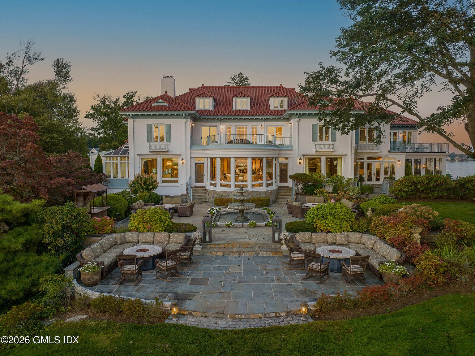 This grand, multi-story estate features a classic Mediterranean-inspired design with a red tile roof and crisp white stucco exterior. The rear of the home opens onto a sprawling stone terrace, complete with a central tiered fountain, symmetrical seating areas with fire pits, and lush, manicured landscaping. The perspective is captured from an elevated vantage point, showcasing the home's impressive scale, architectural symmetry, and its scenic waterfront setting at dusk.