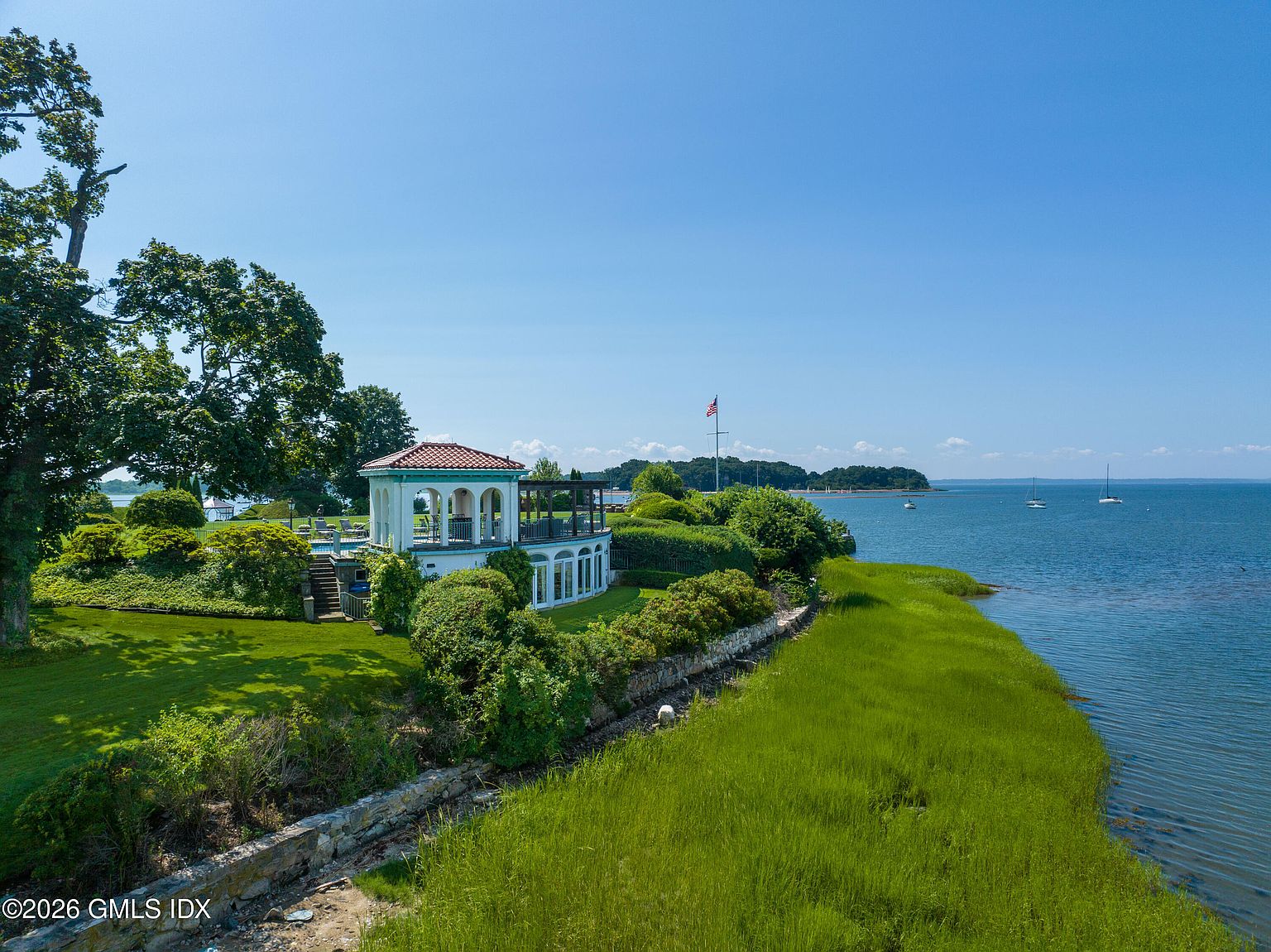 This elevated aerial perspective captures a luxurious waterfront estate featuring a distinctive Mediterranean-style pavilion with a red-tiled roof and arched openings. The property is surrounded by lush green lawns, manicured landscaping, and a stone retaining wall that separates the grounds from the coastal marshland and open water. A tall flagpole stands prominently on the property, which overlooks a serene bay dotted with distant sailboats under a clear blue sky.
