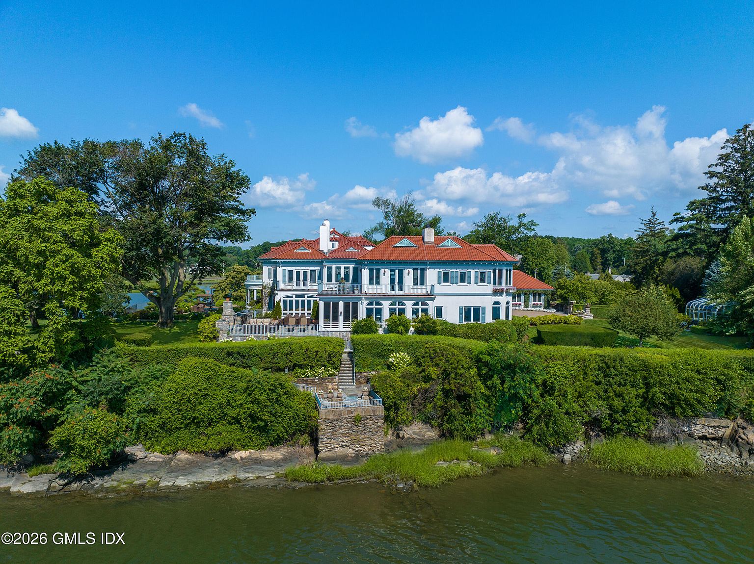 This aerial perspective captures a grand, multi-story estate situated directly on a waterfront. The home features a classic architectural style with a red-tiled roof, white exterior walls, and numerous windows overlooking the water, complemented by a manicured hedge-lined terrace and a stone staircase leading down to the shoreline. The lush green landscaping and expansive blue sky create a serene and prestigious atmosphere for this waterfront property.