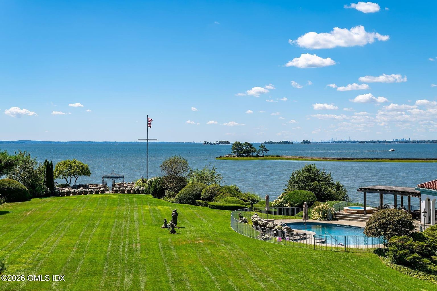 This expansive waterfront property features a meticulously manicured lawn that slopes gently toward a scenic bay. The outdoor space includes a private swimming pool enclosed by a metal fence, a pergola-covered patio area, and lush, mature landscaping. The elevated perspective captures a breathtaking panoramic view of the water, distant islands, and a clear blue sky, emphasizing the property's serene and luxurious coastal setting.