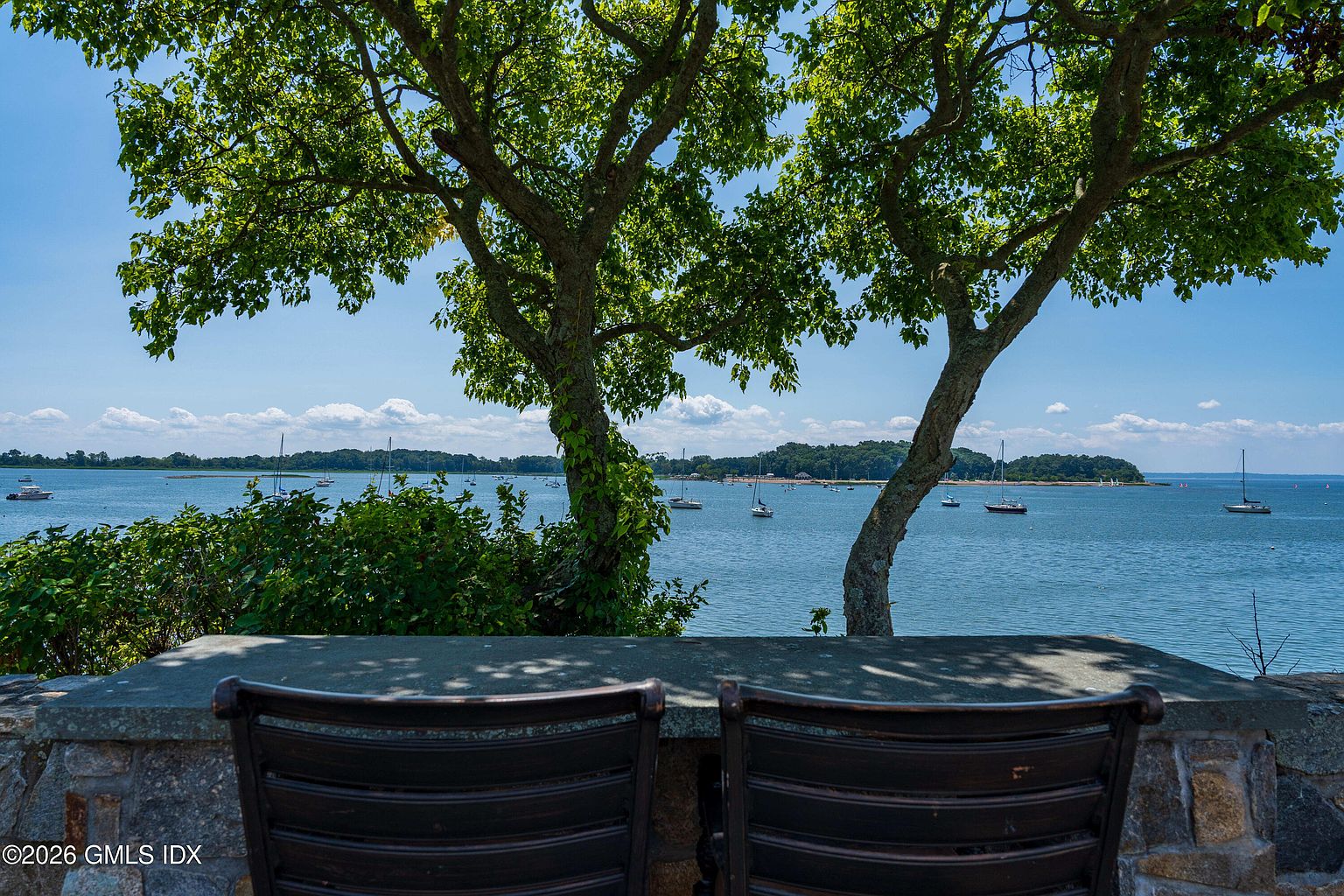 This image captures a serene outdoor seating area overlooking a tranquil bay filled with sailboats. Two dark metal chairs are positioned against a stone-topped wall, framed by the lush green canopy of two mature trees that provide a natural focal point. The perspective is from a seated position, emphasizing the peaceful, scenic waterfront view that defines this property's outdoor living space.